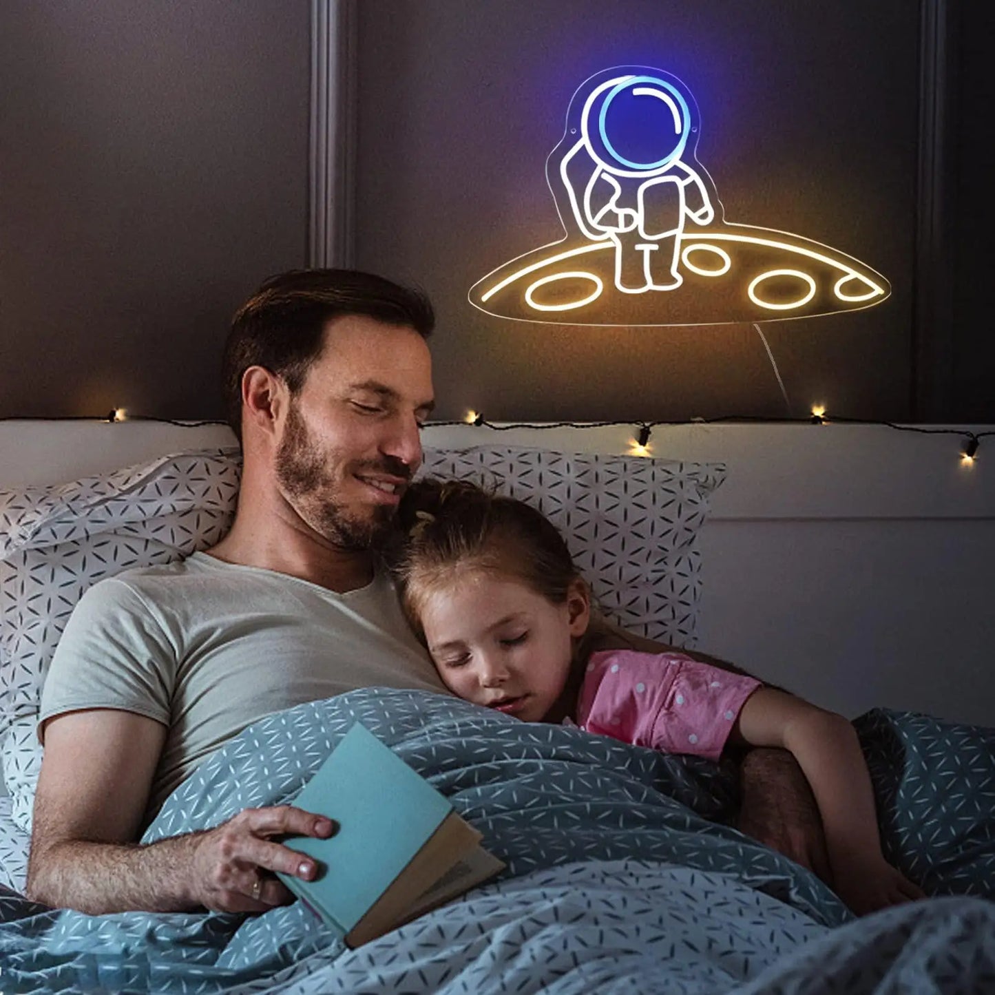 Man reading a book to a child in bed with a neon sign of an astronaut on a spaceship above them.