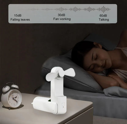 White fan on a nightstand next to a clock, with a woman sleeping in the background.