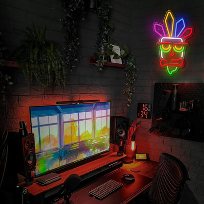 Neon sign of a mask on a dark wall above a computer setup with a colorful screen.